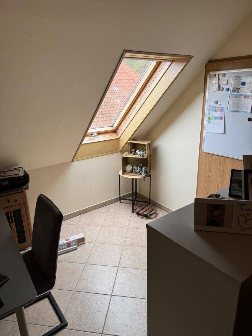 Dachzimmer mit großem Dachfenster, Fliesenboden und einfacher Einrichtung.