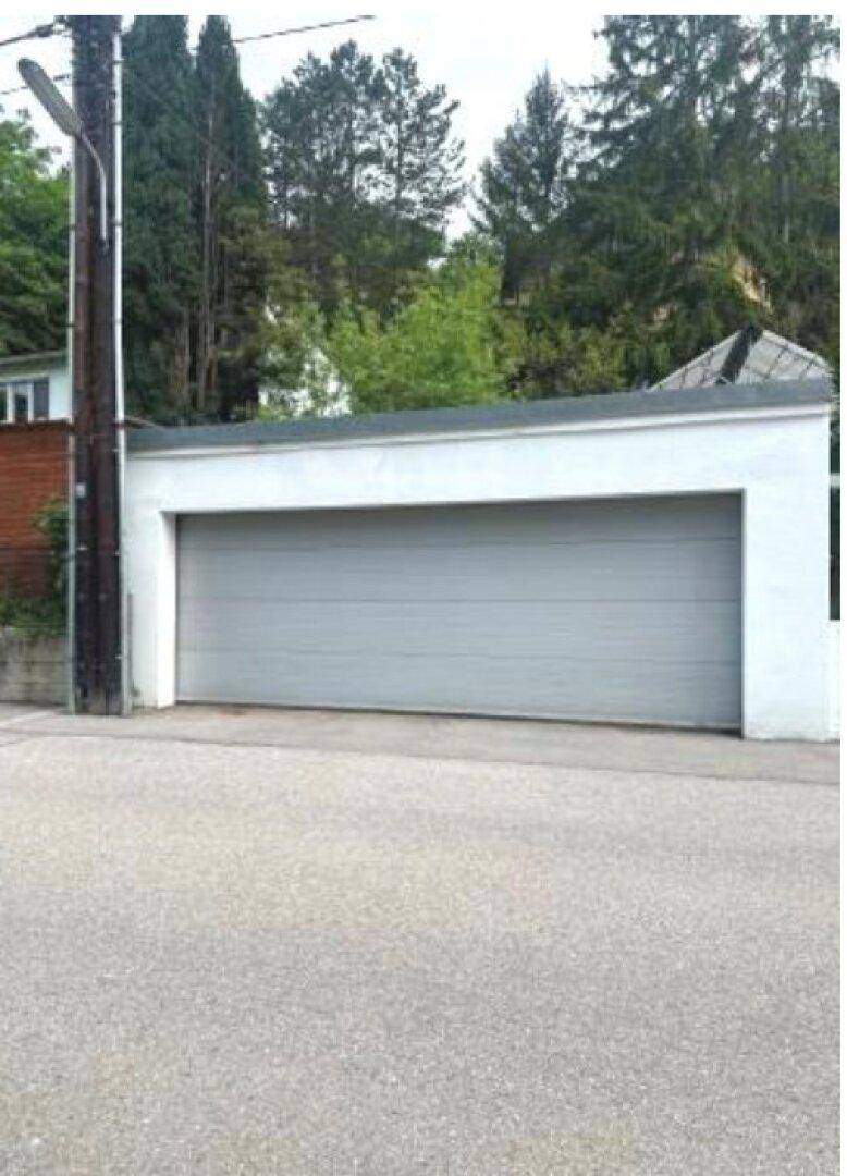 Eine weiße Garage mit grauem Tor, von der Straße aus gesehen, umgeben von Bäumen und einem Strommast.