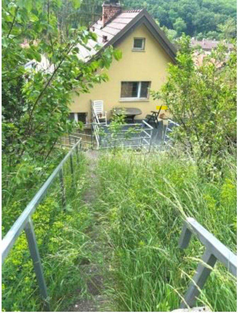 Blick von einer Metalltreppe auf ein gelbes Haus mit einer möblierten Terrasse, umgeben von üppigem Grün.