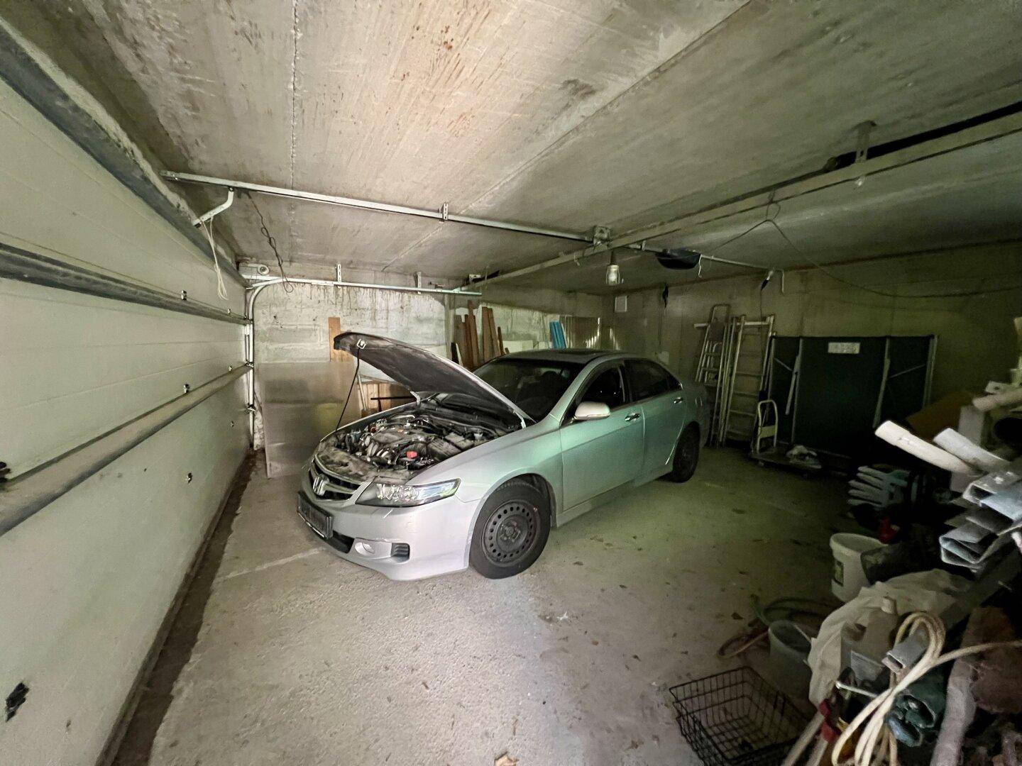 Unaufgeräumte Garage mit einem Auto, Werkzeugen und verschiedenen Gegenständen an Betonwänden.