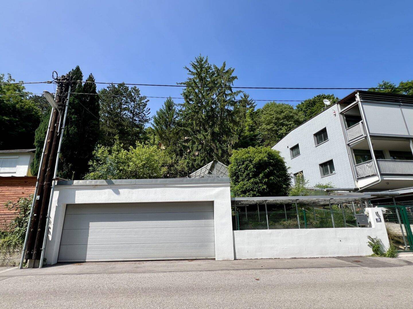 Weiße Garage mit grauem Tor neben einem Mehrfamilienhaus und einem bewaldeten Hang.