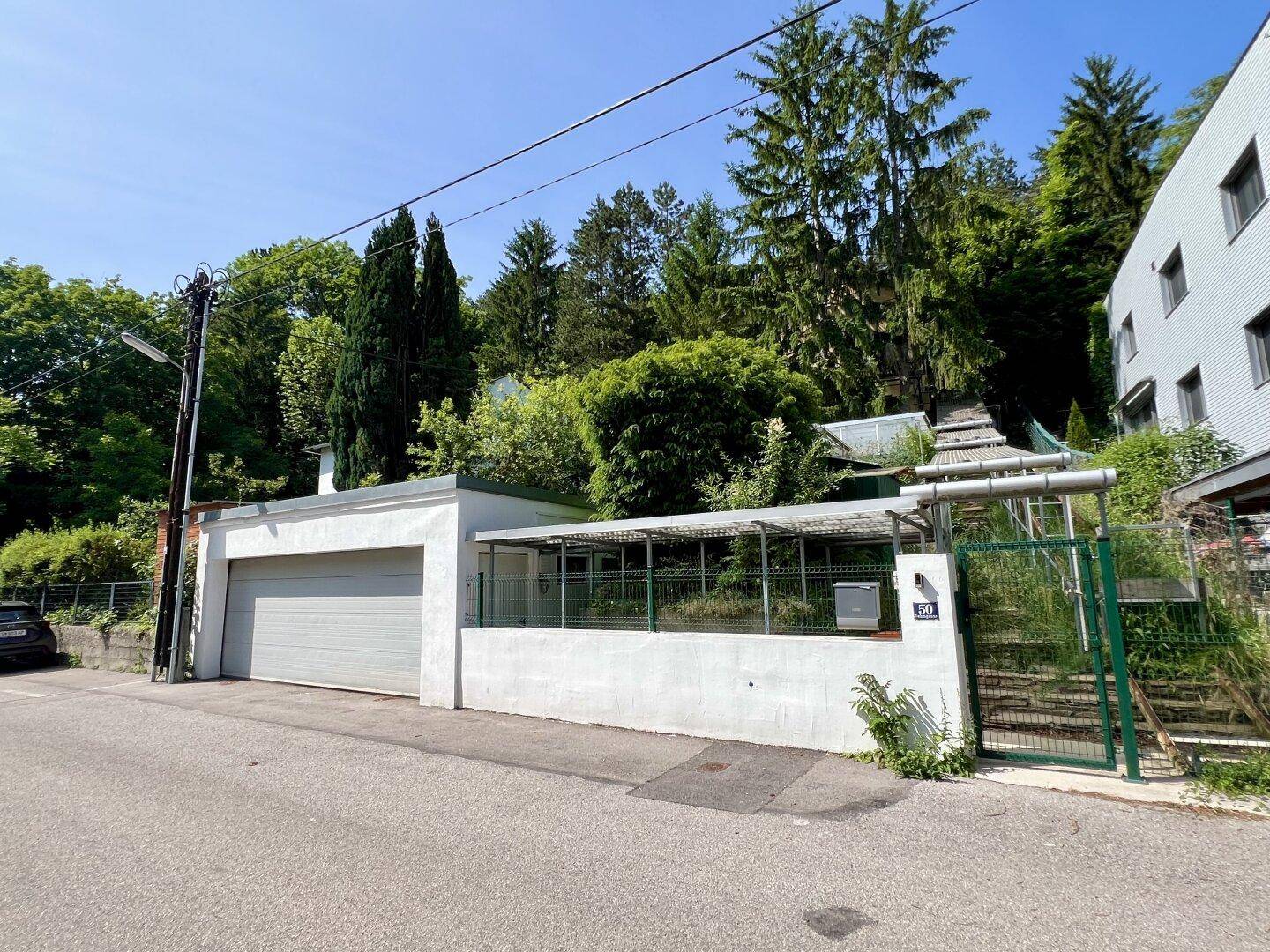 Weitläufige Außenansicht mit weißer Garage, Zaun und einem Gebäude am bewaldeten Hang.