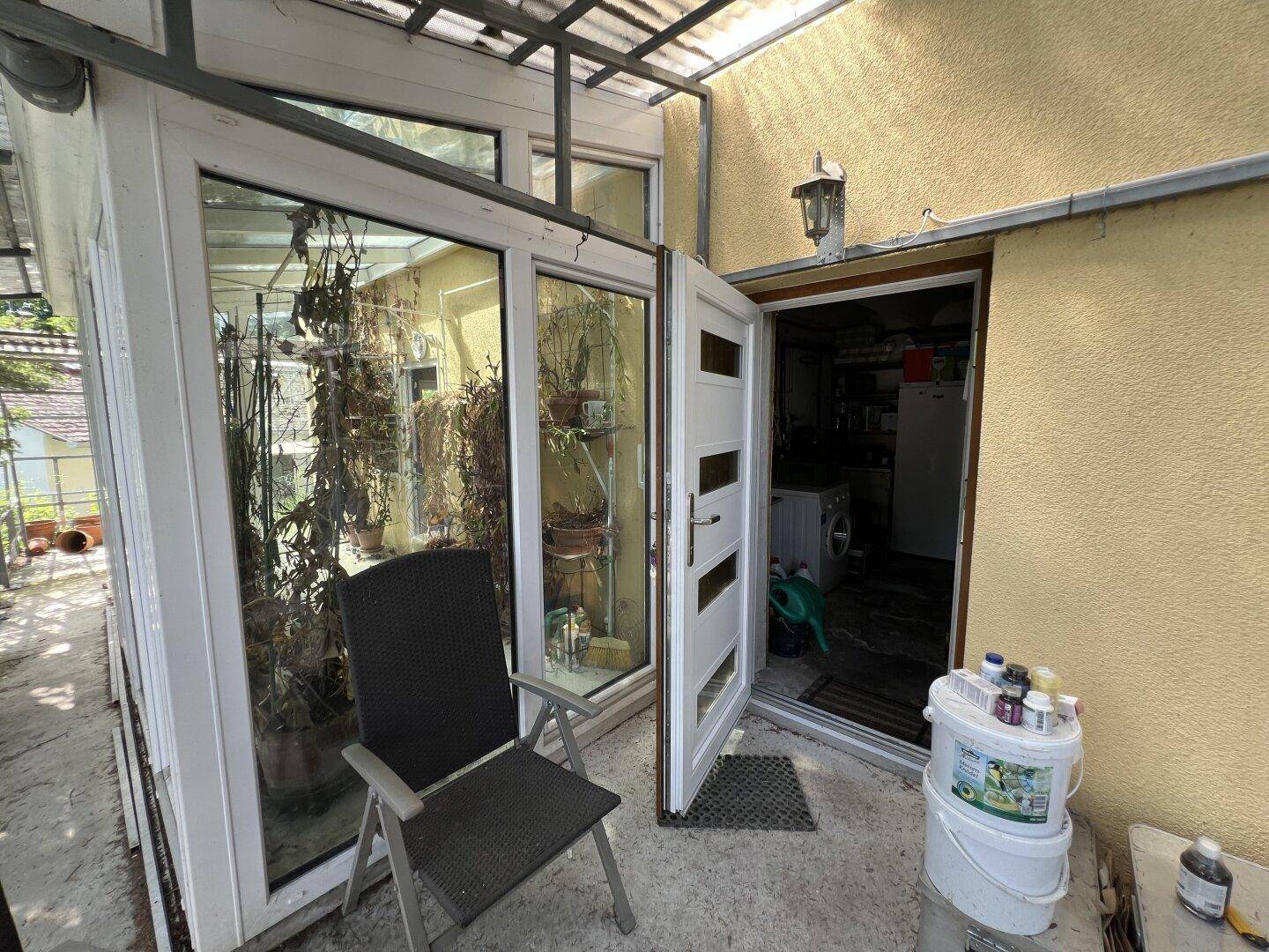 Eingangsbereich eines gelben Hauses mit weißer Tür neben einem verglasten Wintergarten und einem Stuhl.