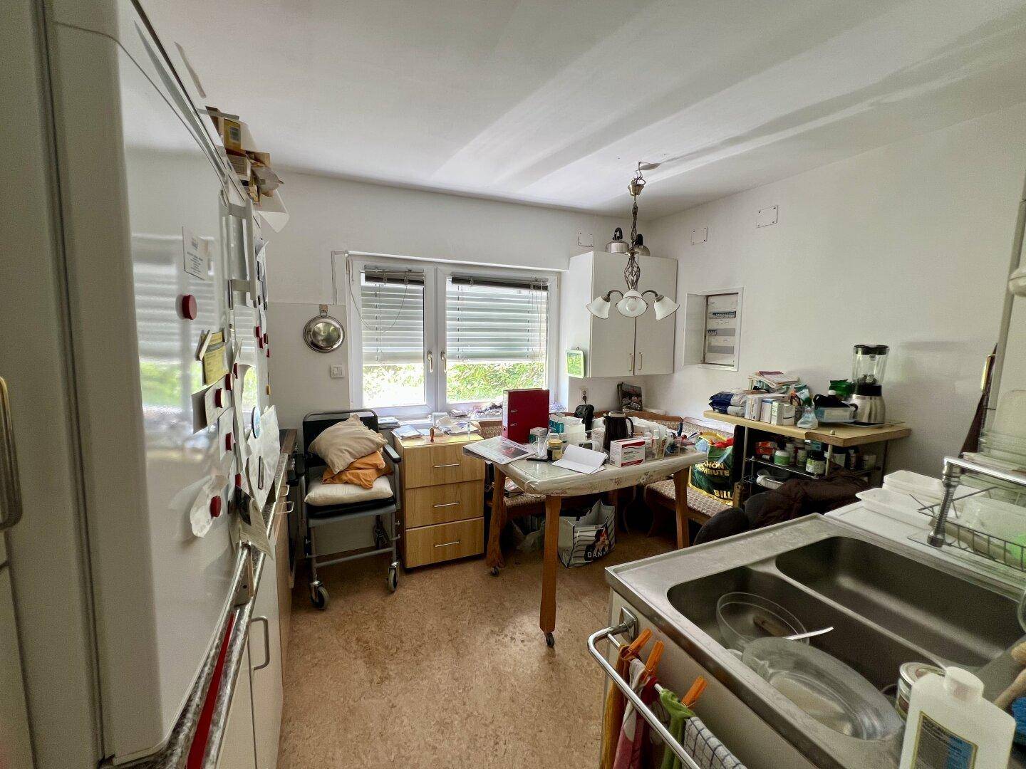 Küche mit Esstisch, Spüle, Kühlschrank und Fenster mit Blick ins Grüne, wirkt unaufgeräumt.