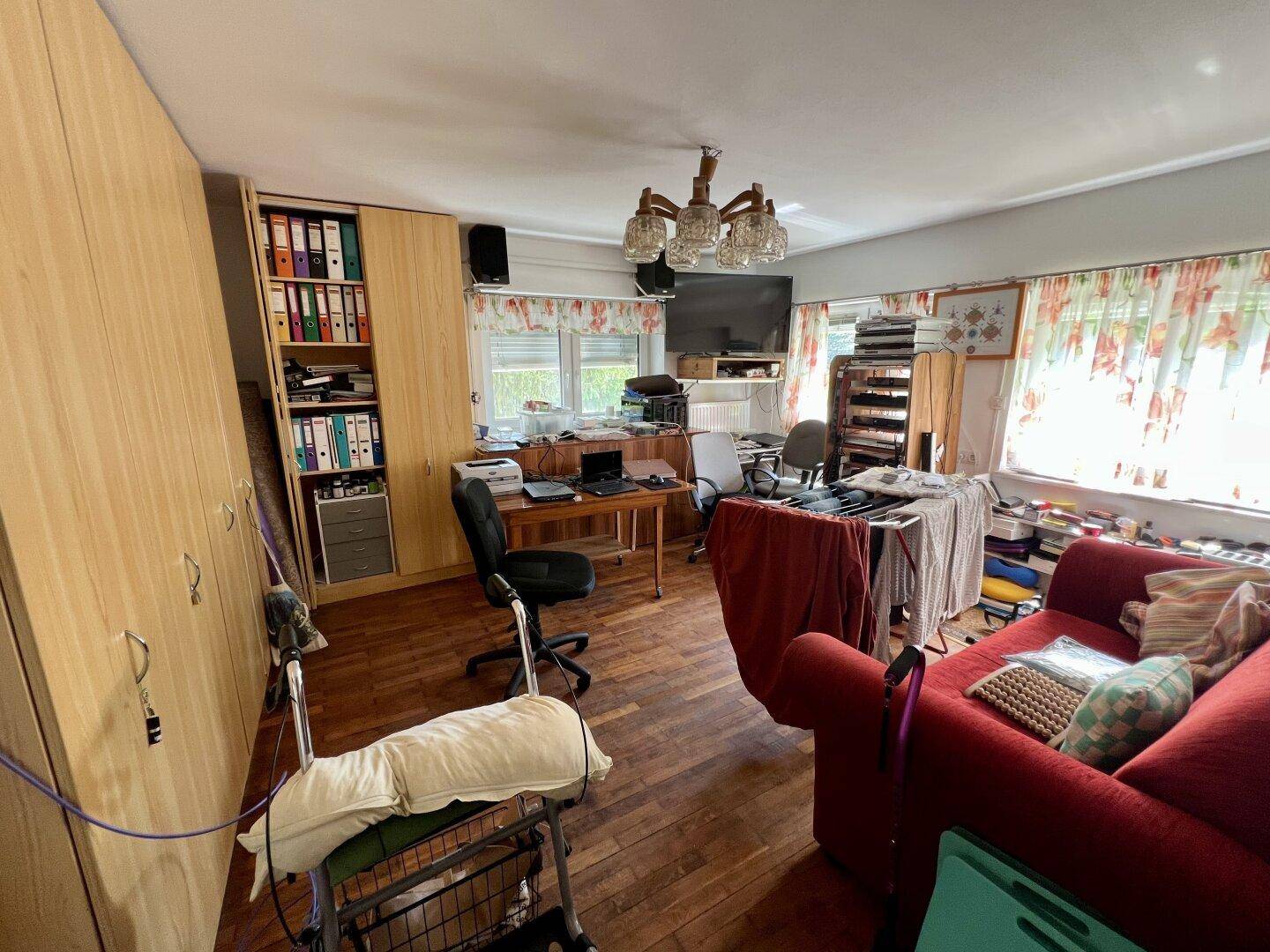 Wohn- oder Arbeitszimmer mit rotem Sofa, Schreibtisch, Stühlen und Holzschränken, mit Fenstern.
