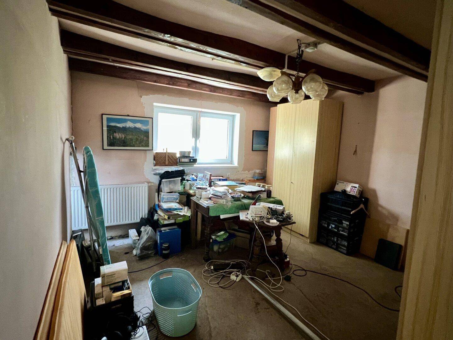 Raum mit sichtbaren Holzbalken, Fenster, Schreibtisch und verschiedenen Gegenständen, als Büro genutzt.