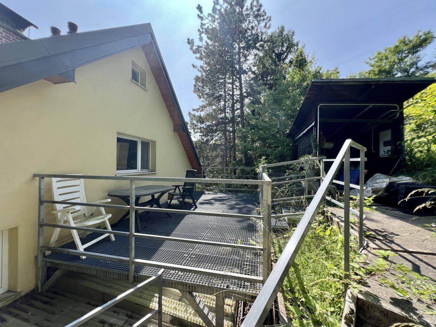 Außenterrasse mit Metallgitterboden und Geländer neben einem gelben Haus, mit Schuppen und Bäumen.