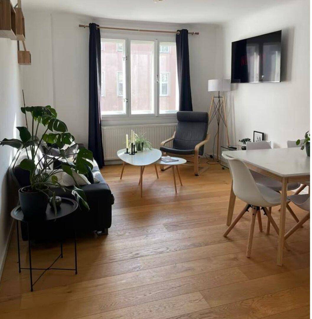 Offener Wohn-Essbereich mit Holzboden, dunklem Sofa und Esstisch, beleuchtet durch große Fenster.