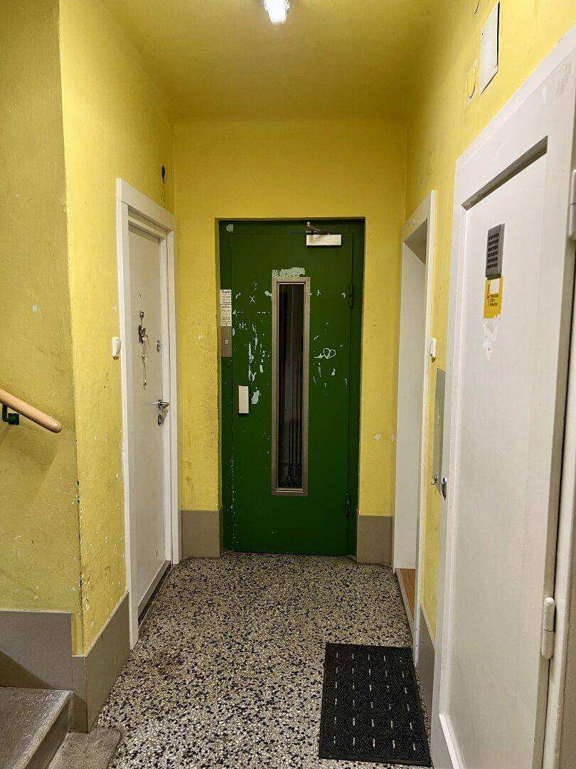 Gelber Hausflur mit Terrazzoboden und einer grünen Aufzugstür im Mehrfamilienhaus.