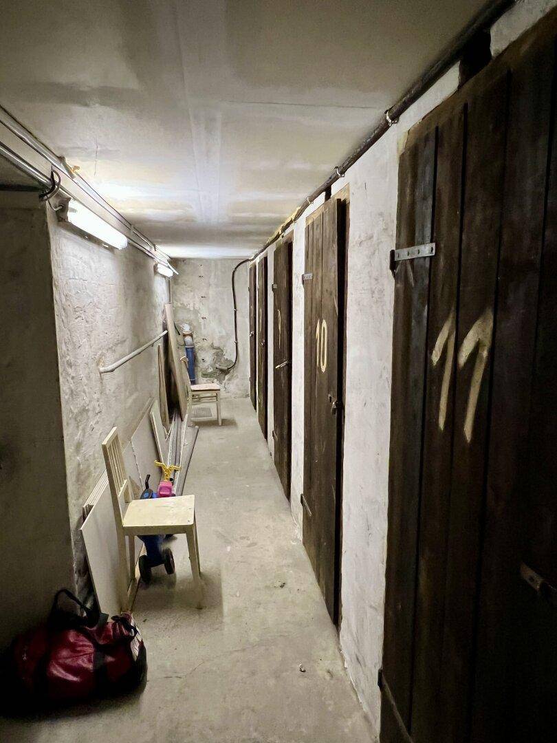 Kellergang mit mehreren Holzabteilen und Betonboden, beleuchtet von Neonröhren.