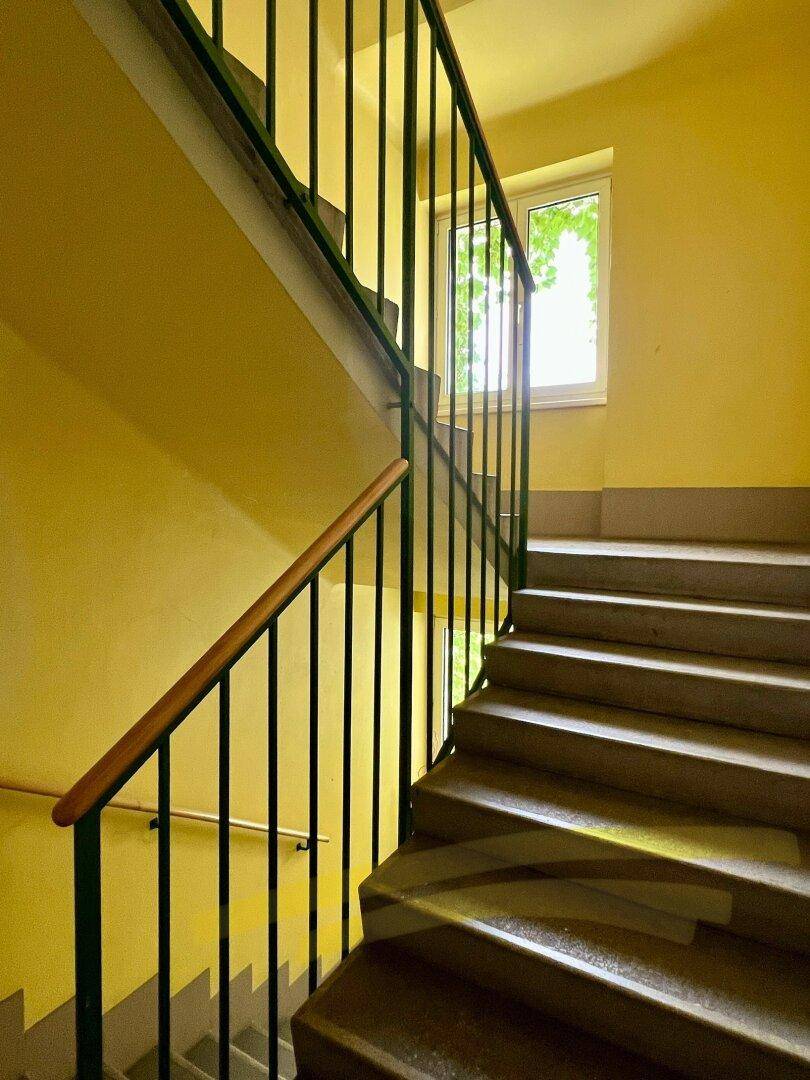 Gelbes Treppenhaus mit Steinstufen, grünem Geländer und Fenster für natürliches Licht.