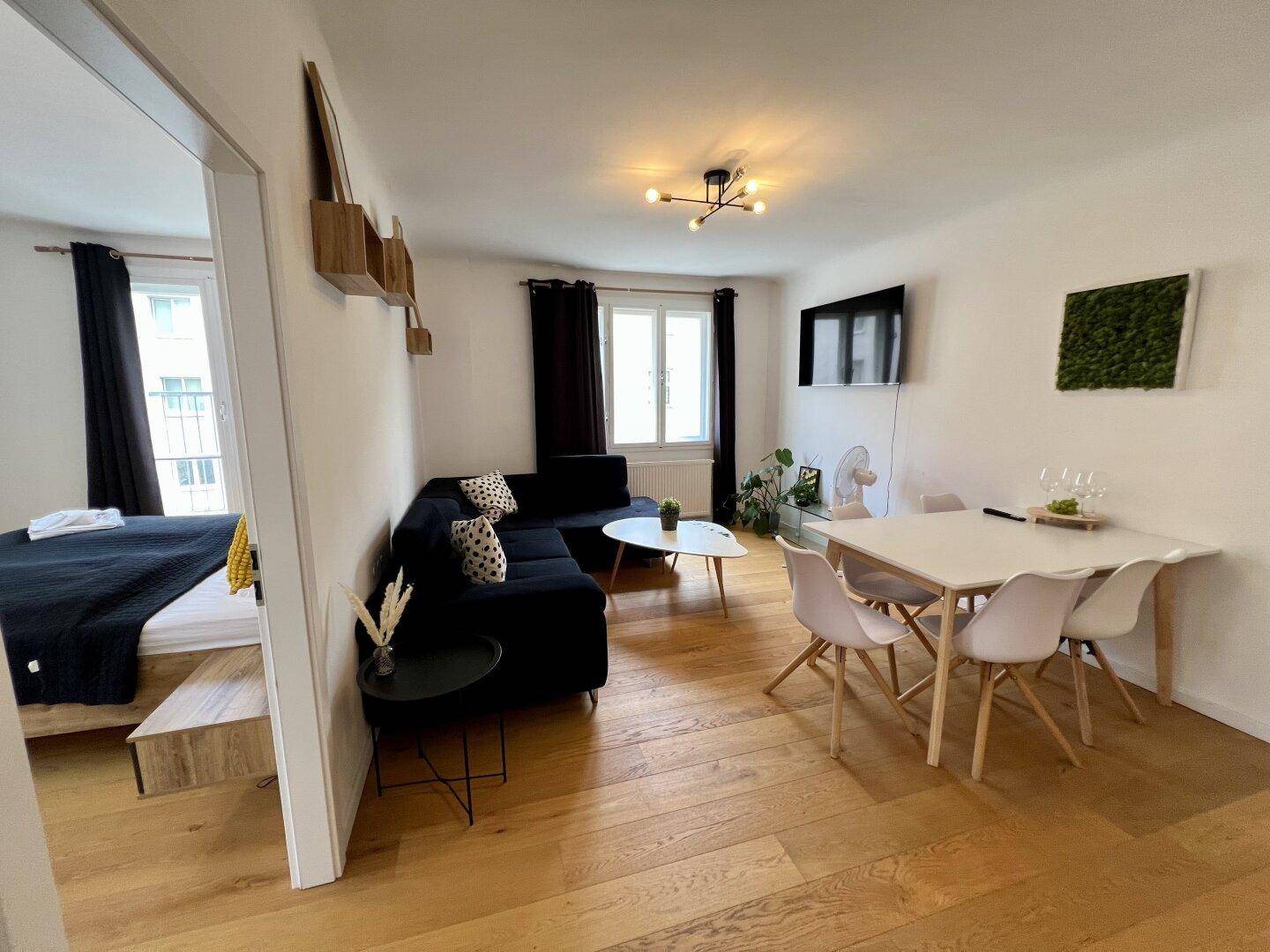 Heller Wohn-Essbereich mit Holzboden, dunklem Sofa und Esstisch, angrenzend an ein Schlafzimmer.