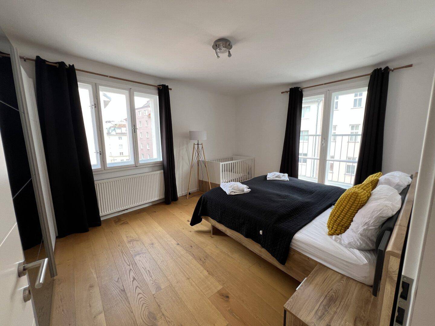 Helles Schlafzimmer mit Holzboden, dunklem Bett und zwei Fenstern mit Blick auf die Umgebung.