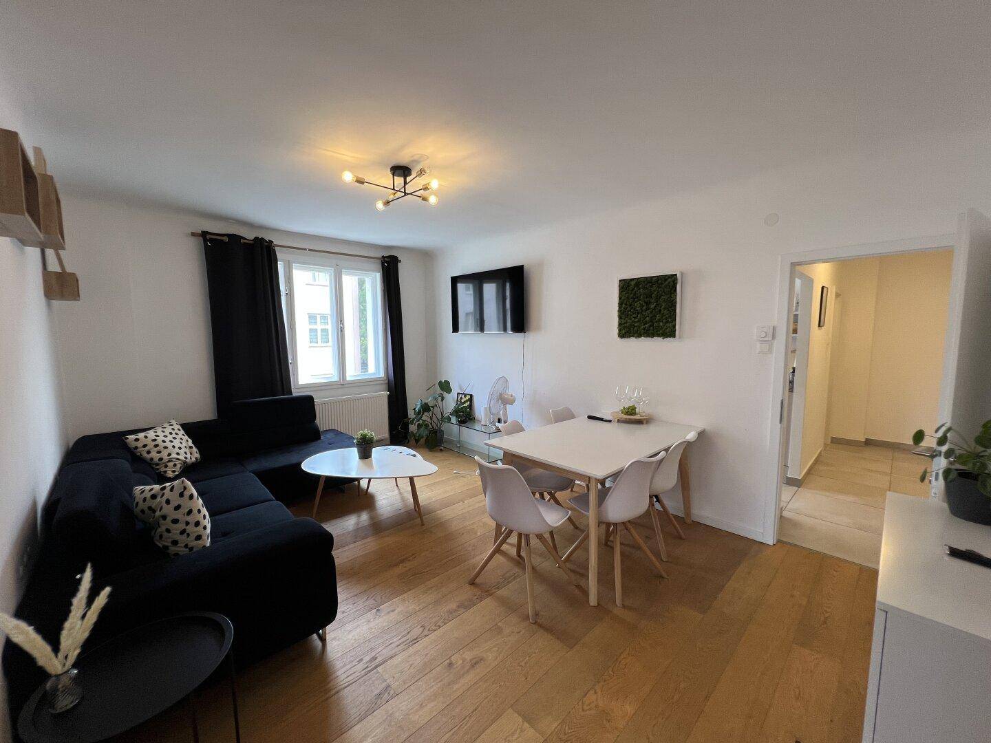Geräumiger Wohn-Essbereich mit Holzboden, dunklem Sofa und modernem Esstisch für vier Personen.