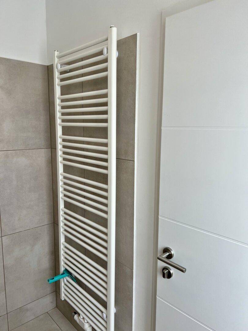 Moderner Handtuchheizkörper in einem Badezimmer mit hellen Fliesen und weißer Tür.
