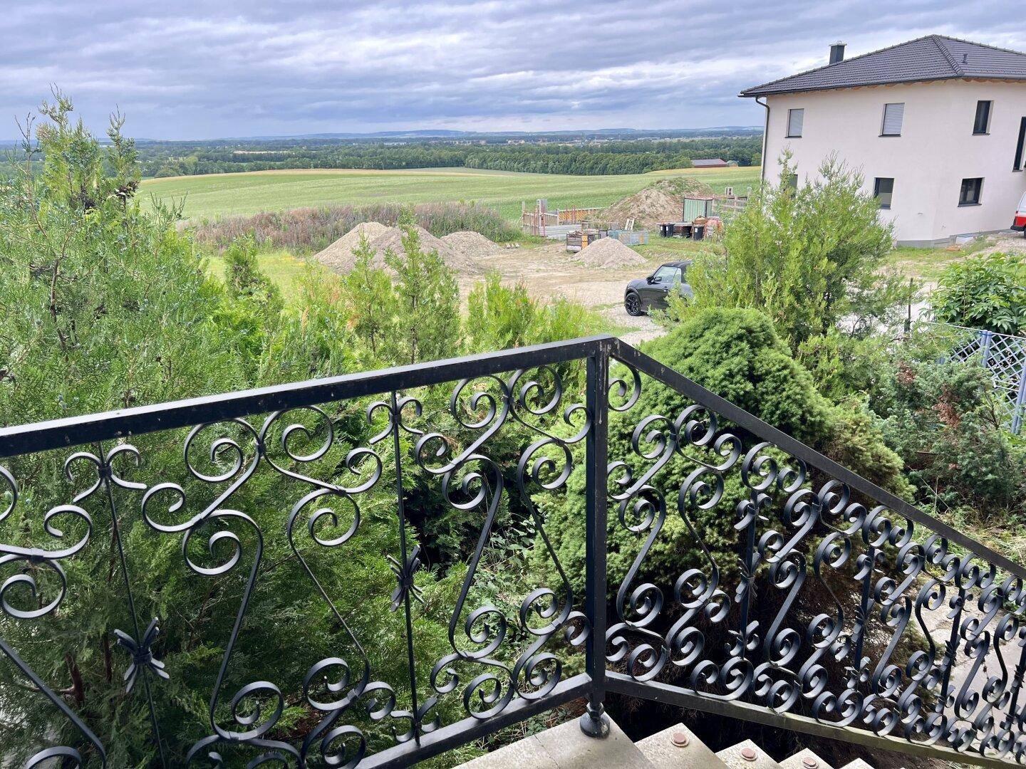 Weitblick über die Landschaft mit Bäumen und einem Haus, gesehen von einem Balkon mit schmiedeeisernem Geländer.