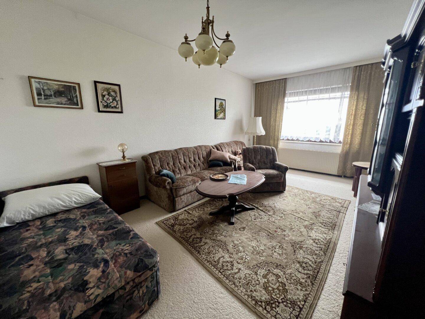 Wohnzimmer mit Sofa, Einzelbett und Teppichboden, hell durch ein großes Fenster.