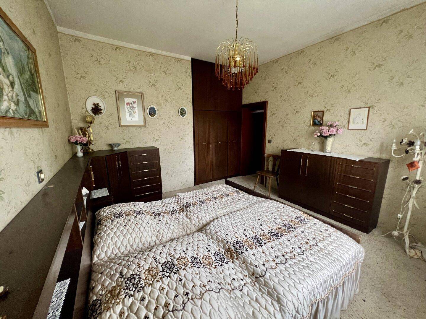 Schlafzimmer mit gemusterter Tapete, dunklen Holzmöbeln und einem klassischen Kronleuchter.