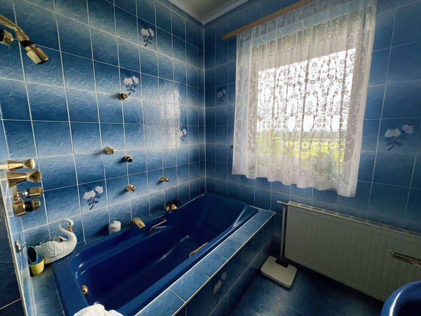 Badezimmer mit blauen Fliesen, Badewanne und Fenster mit weißer Gardine.