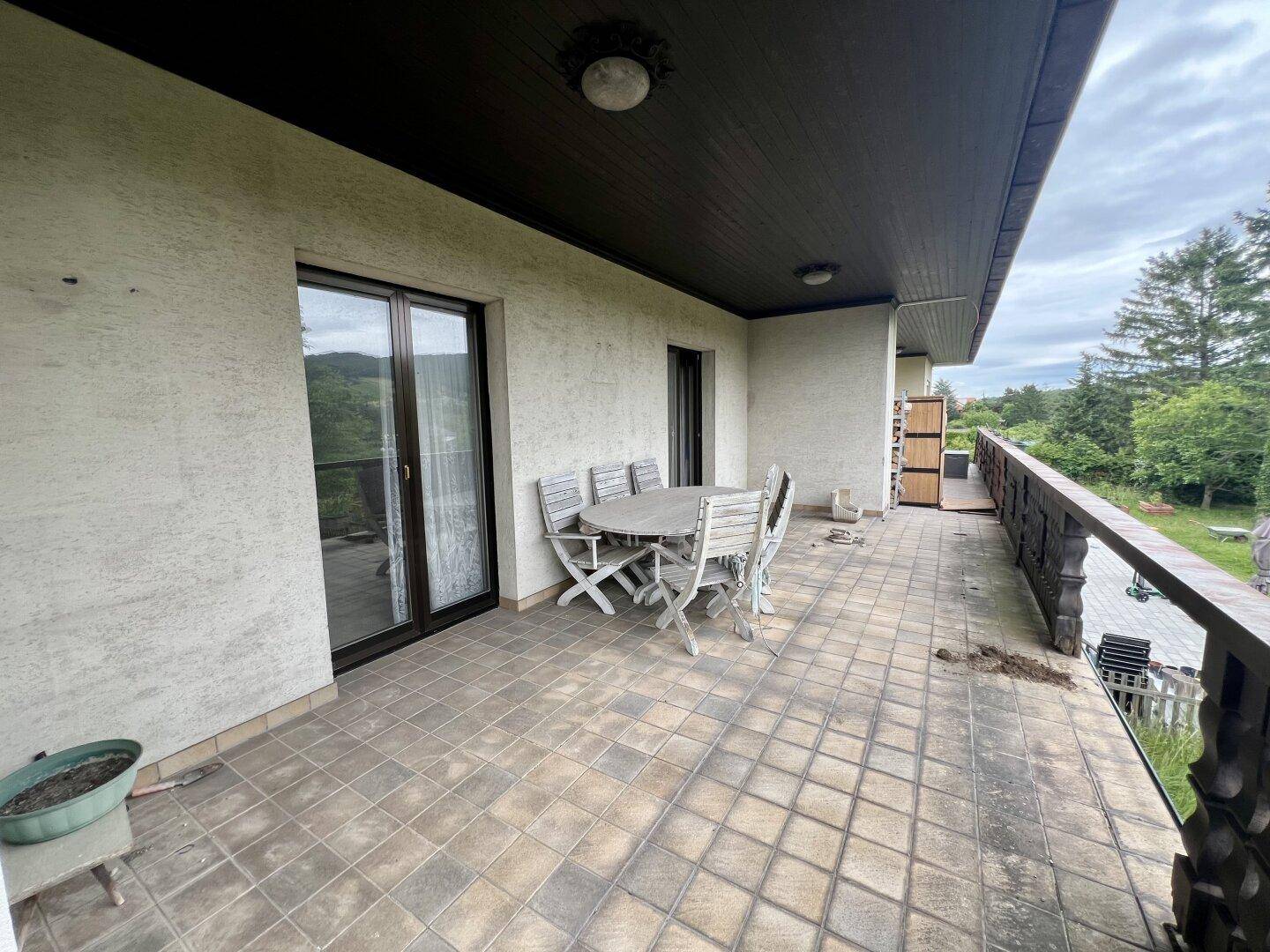 Große geflieste Terrasse mit Holztisch und Stühlen, umgeben von viel Grün.