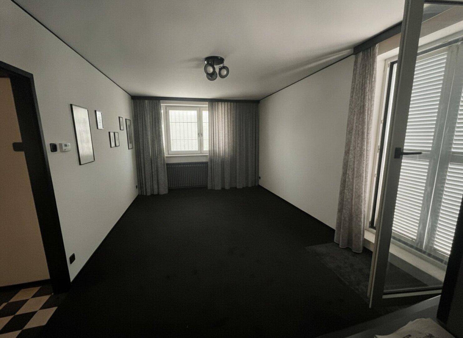 Helles Wohnzimmer mit dunklem Teppichboden, weißen Wänden und Fenstern mit Jalousien.