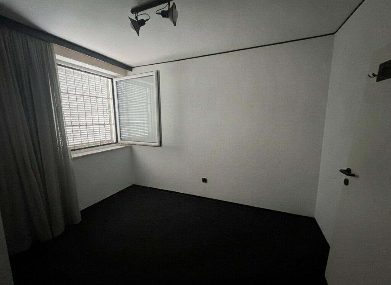 Schlafzimmer mit dunklem Teppichboden, weißen Wänden und einem geöffneten Fenster mit Jalousien.