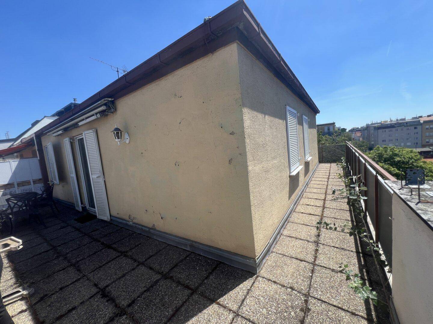Geräumige Terrasse mit gefliestem Boden und einer Hauswand mit Fensterläden unter blauem Himmel.