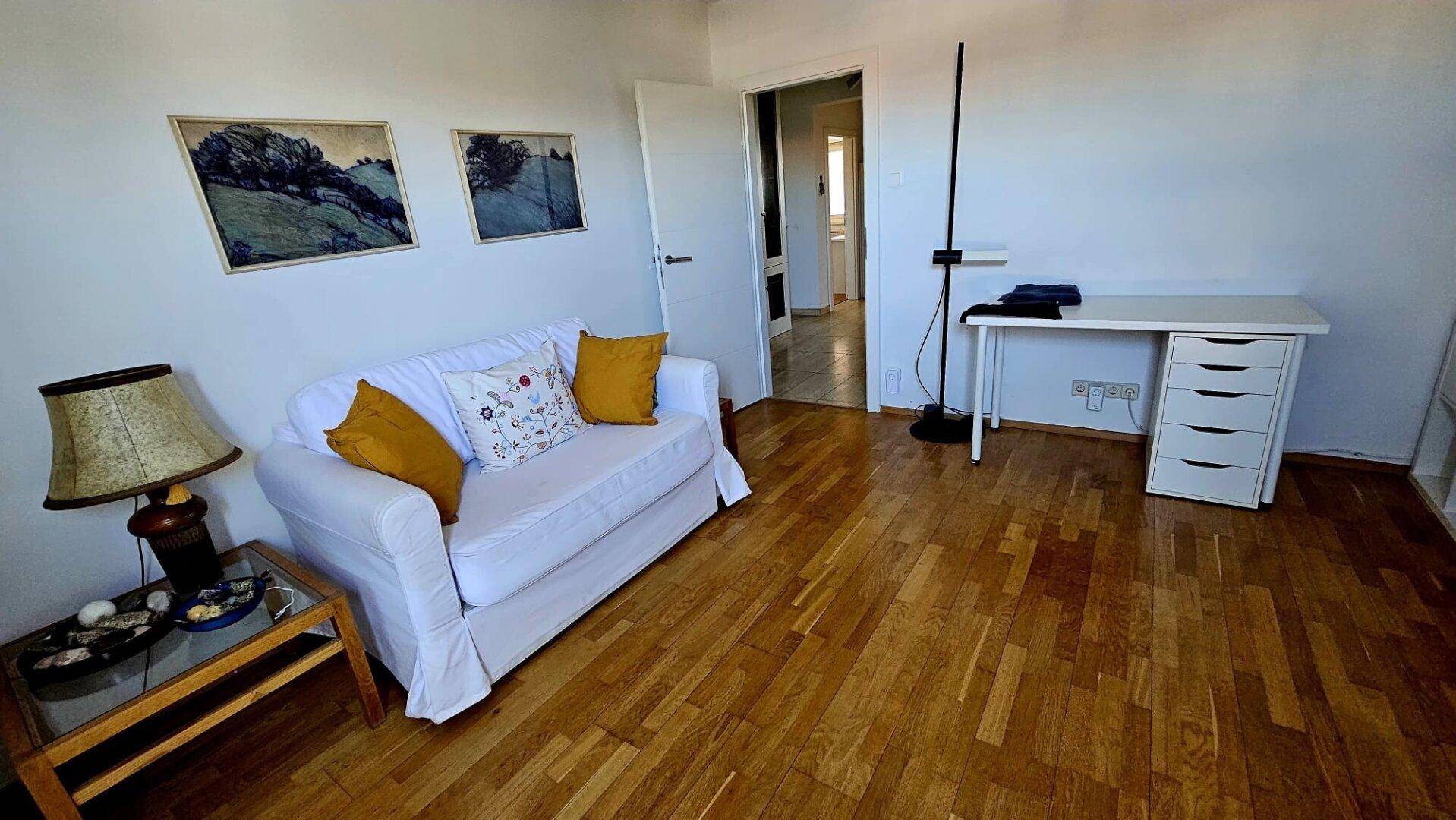Gemütliches Wohnzimmer mit weißem Sofa, Beistelltisch und einem Arbeitsbereich.