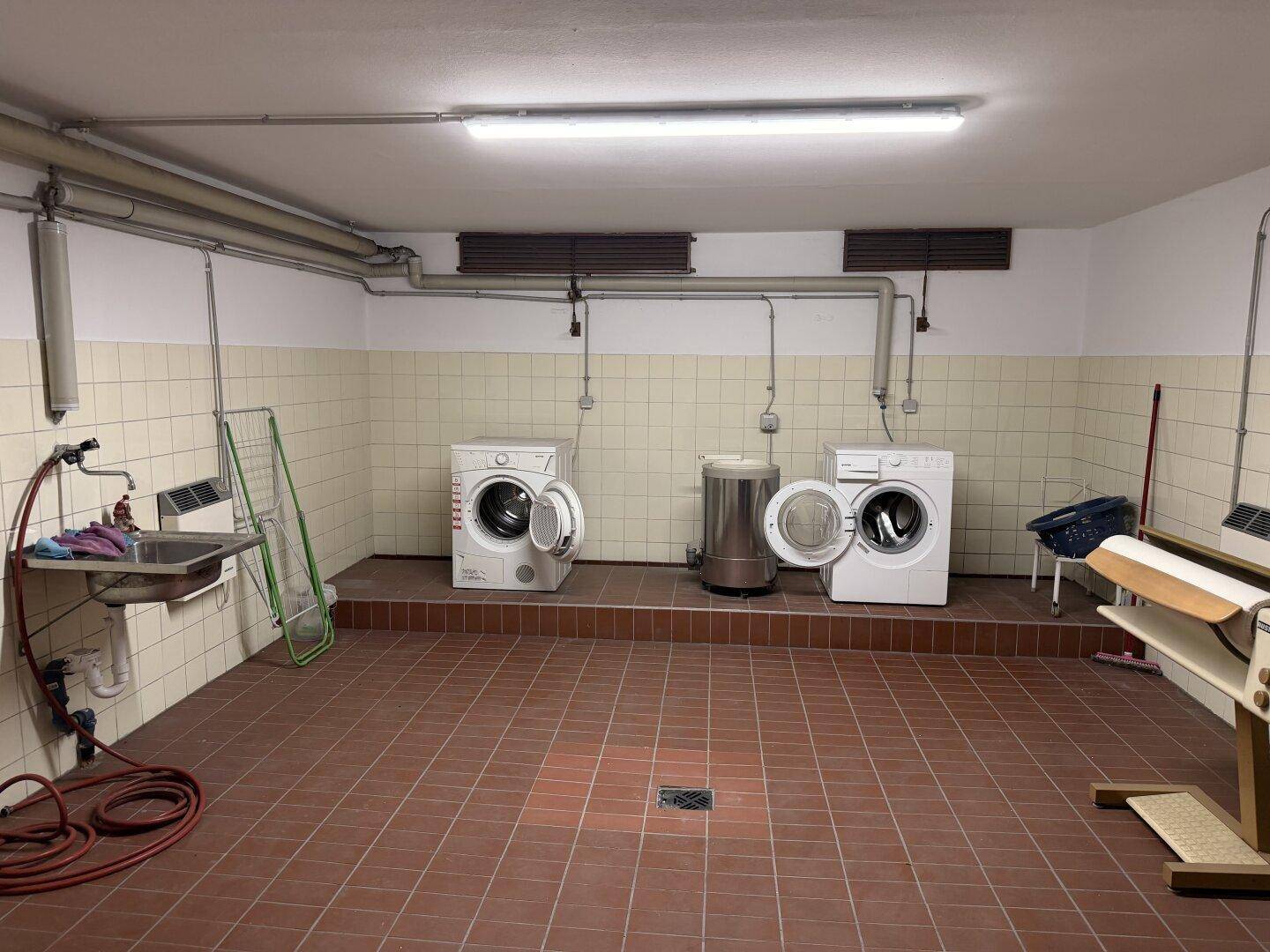 Gemeinschaftlicher Waschraum mit Fliesenboden, Waschmaschinen und einem Waschbecken.