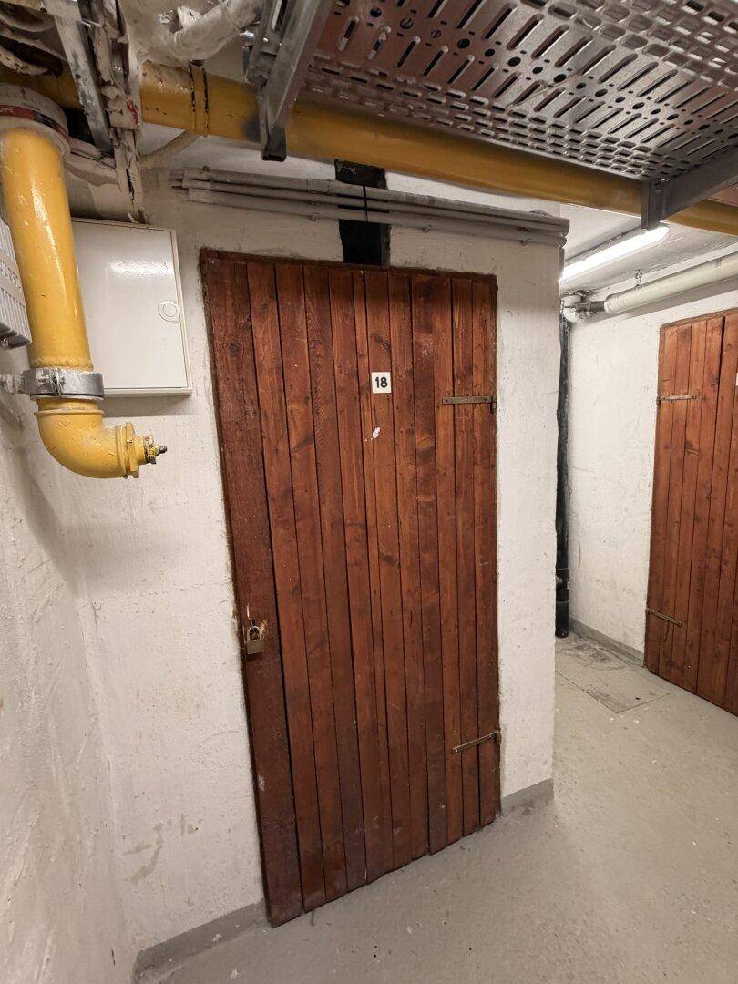 Holztür Nummer 18 in einem Kellerflur mit sichtbaren Rohren und Kabelkanälen an der Decke.