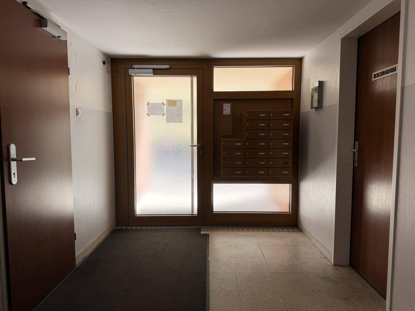 Eingangsbereich eines Mehrfamilienhauses mit Haustür, Briefkästen und Fliesenboden.