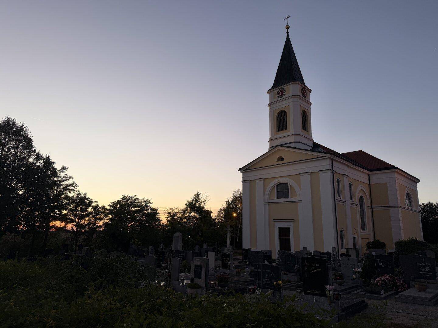 Historische Kirche mit Friedhof bei Sonnenuntergang, umgeben von Bäumen und Grabsteinen.
