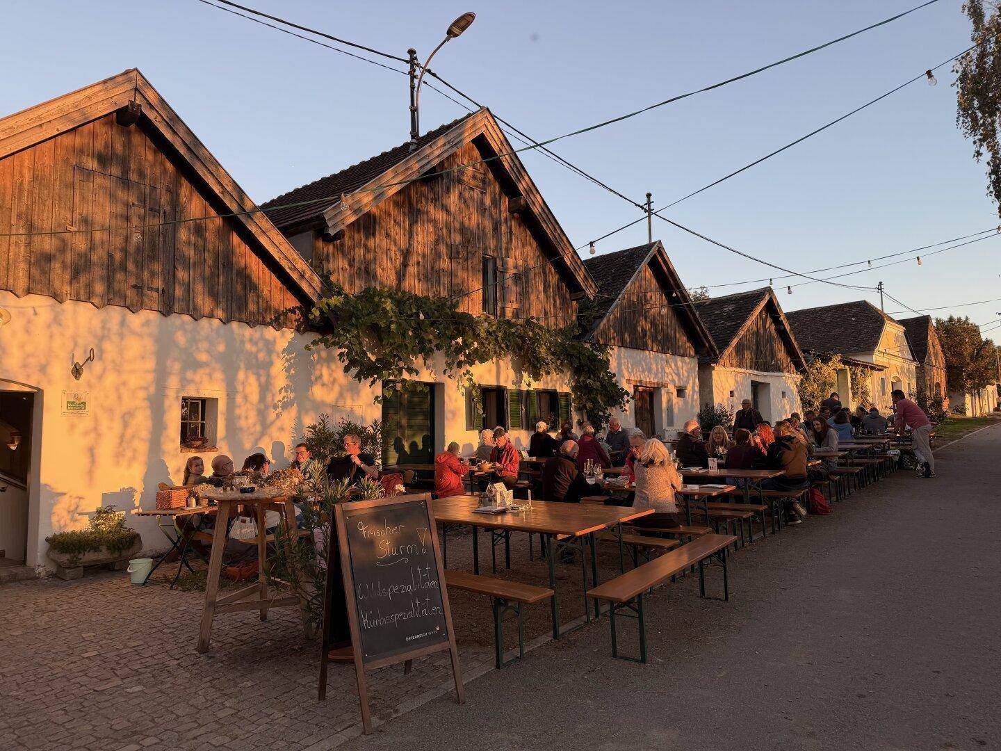 Gemütlicher Außenbereich mit Holztischen und Bänken vor traditionellen Gebäuden bei Sonnenuntergang.