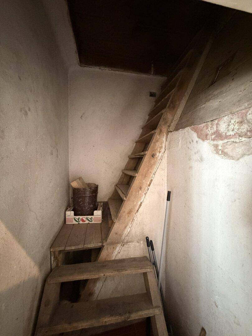 Enger Flur mit steiler Holztreppe, die zu einem oberen Stockwerk führt, in einem alten Gebäude.
