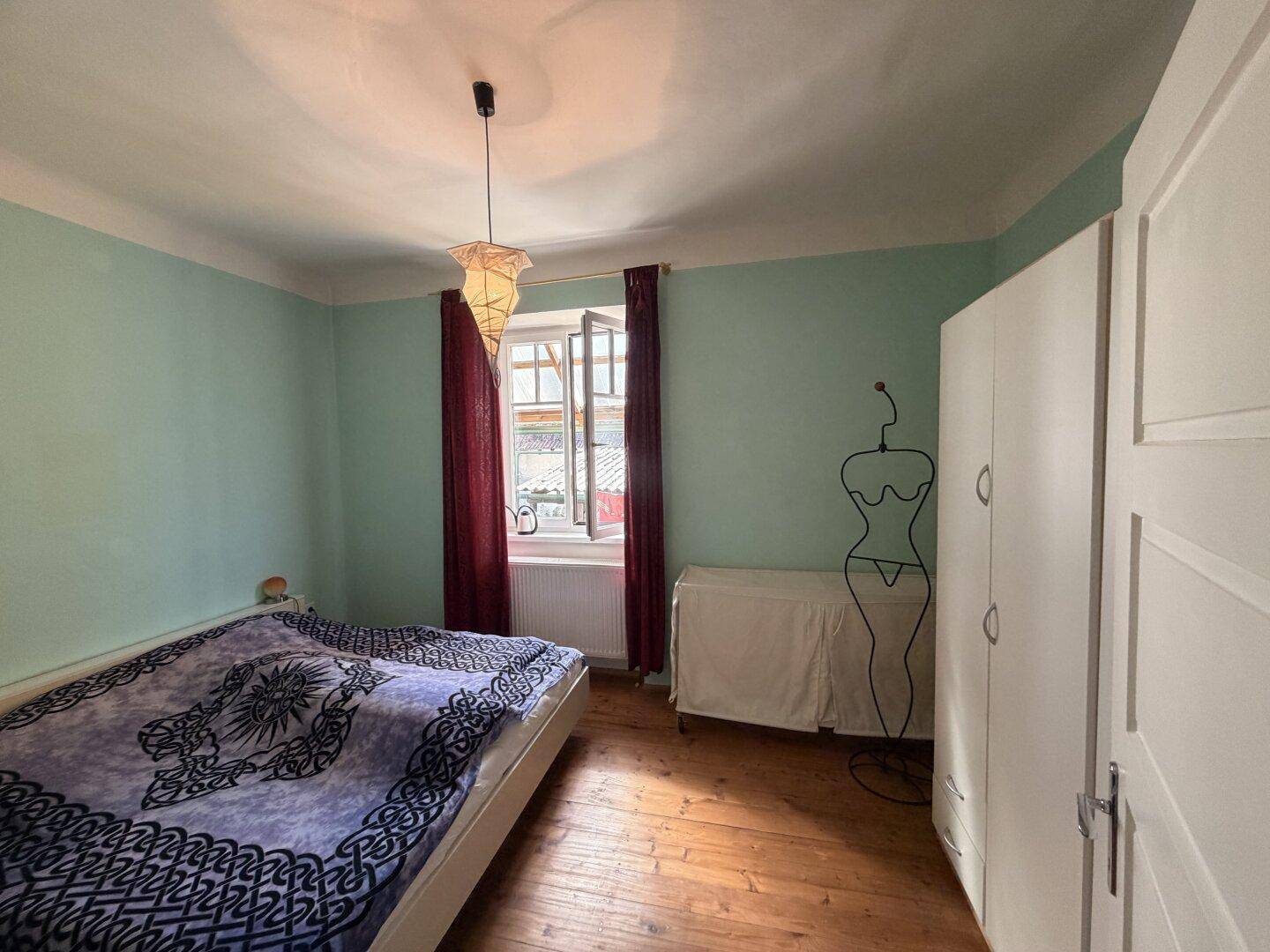 Helles Schlafzimmer mit Holzboden und Fenster mit roten Vorhängen, das viel Tageslicht hereinlässt.