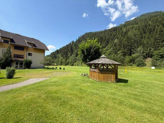 Große grüne Rasenfläche mit Holzpavillon und Blick auf bewaldete Berge unter blauem Himmel.