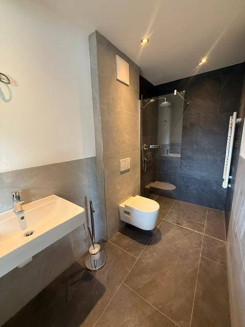 Modernes Badezimmer mit grauen Fliesen, wandhängendem WC und ebenerdiger Dusche.