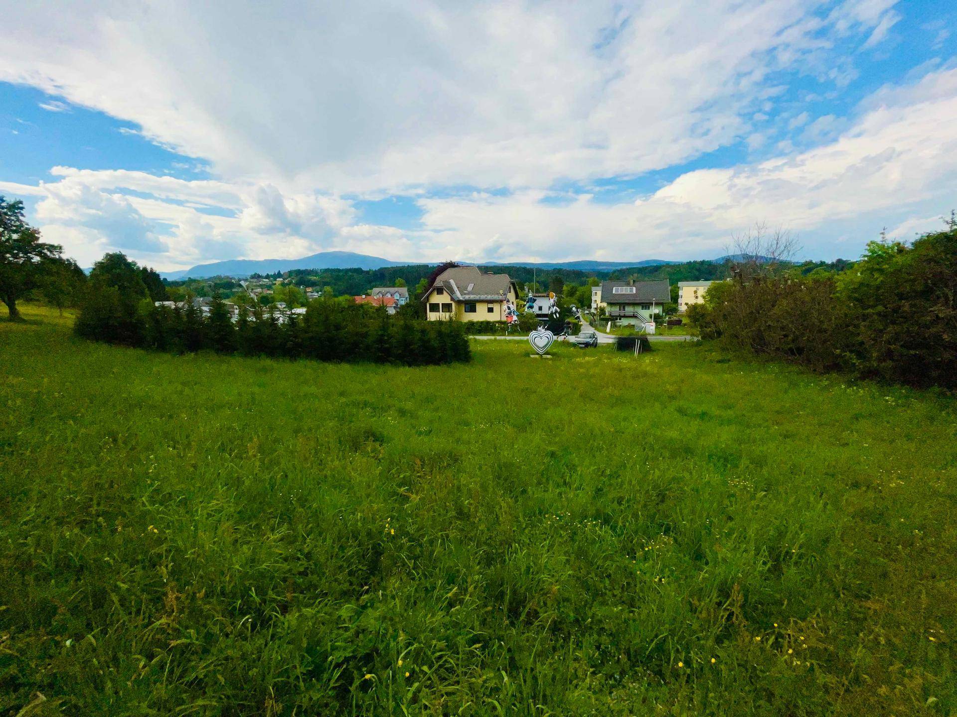 Panoramablick über eine grüne Wiese mit Häusern und einer Bergkette am Horizont.
