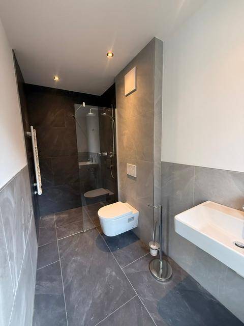 Modernes Badezimmer mit grauen Fliesen, wandhängendem WC und ebenerdiger Dusche.