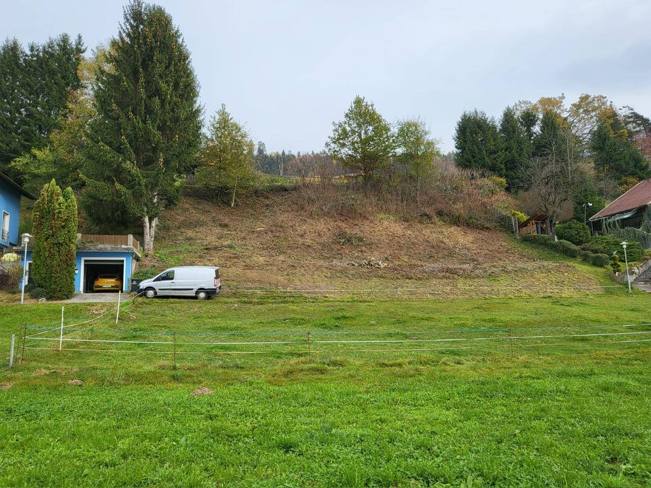 Großes Grundstück mit Rasenfläche, bewaldetem Hang und einer Garage am Rand des Anwesens.