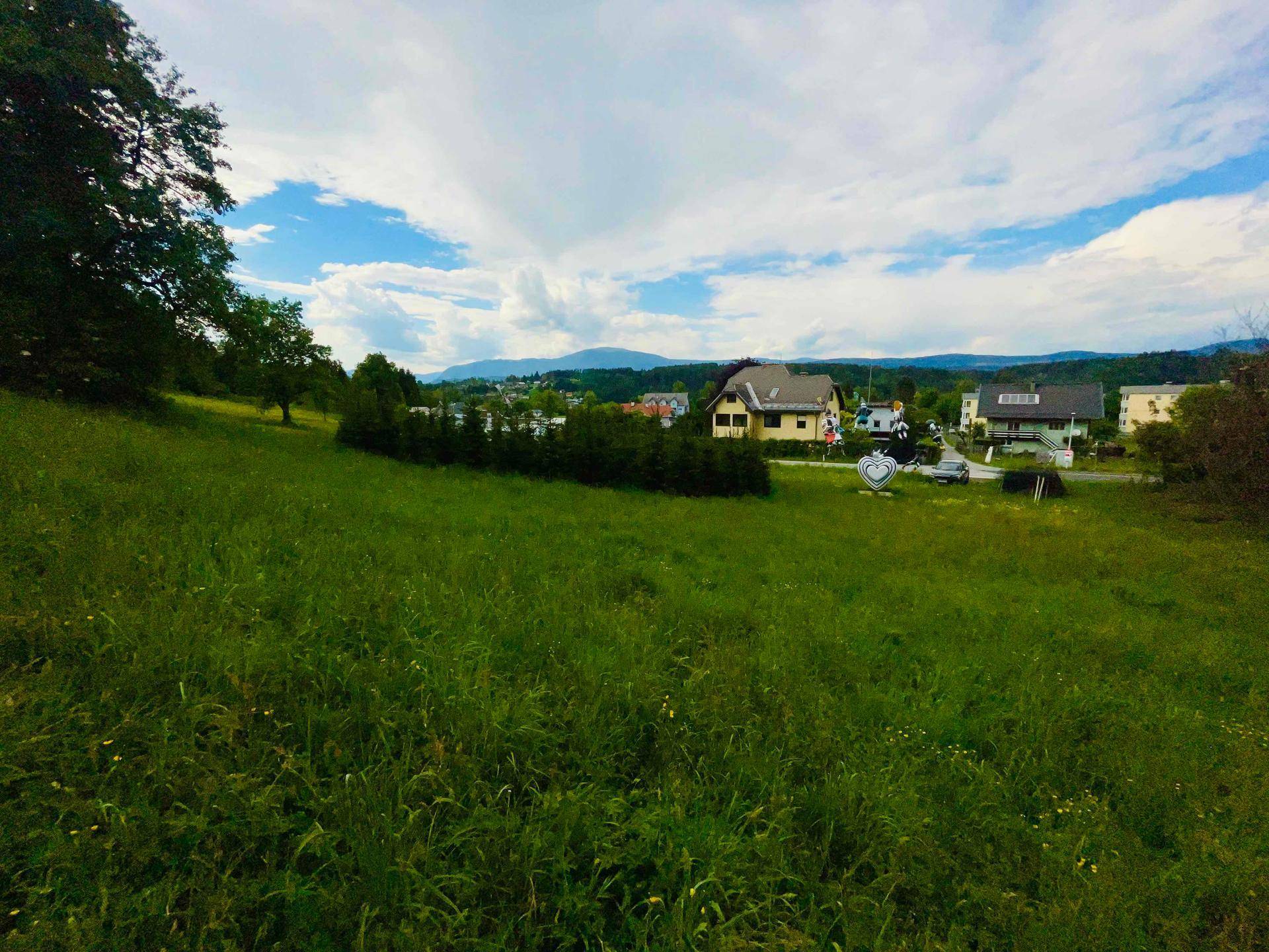 Idyllische grüne Landschaft mit weitläufigen Wiesen, Häusern und Bergen unter einem bewölkten Himmel.