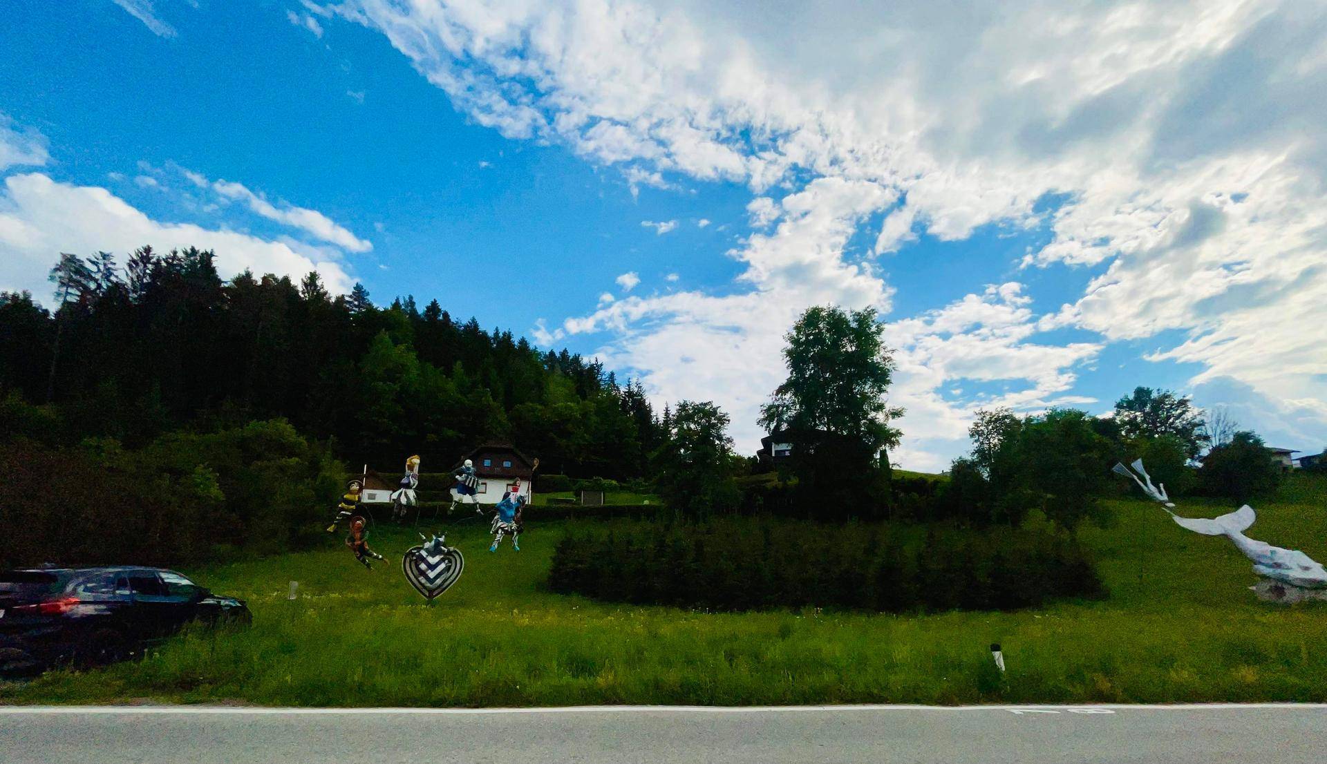 Blick über eine Straße auf eine bewaldete Anhöhe mit Häusern und dekorativen Elementen.