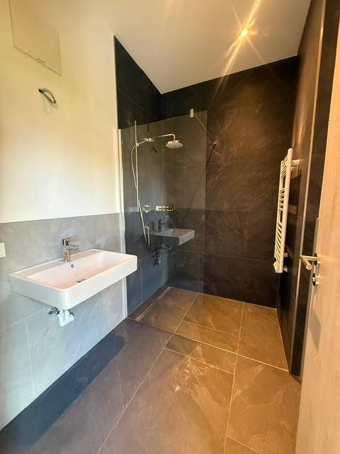 Modernes Badezimmer mit dunklen Fliesen, bodengleicher Dusche, Wandwaschbecken und Handtuchheizkörper.