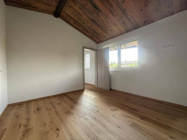 Heller Raum mit Holzboden, schräger Holzdecke und Fenster, bietet Zugang zu einem weiteren Zimmer.