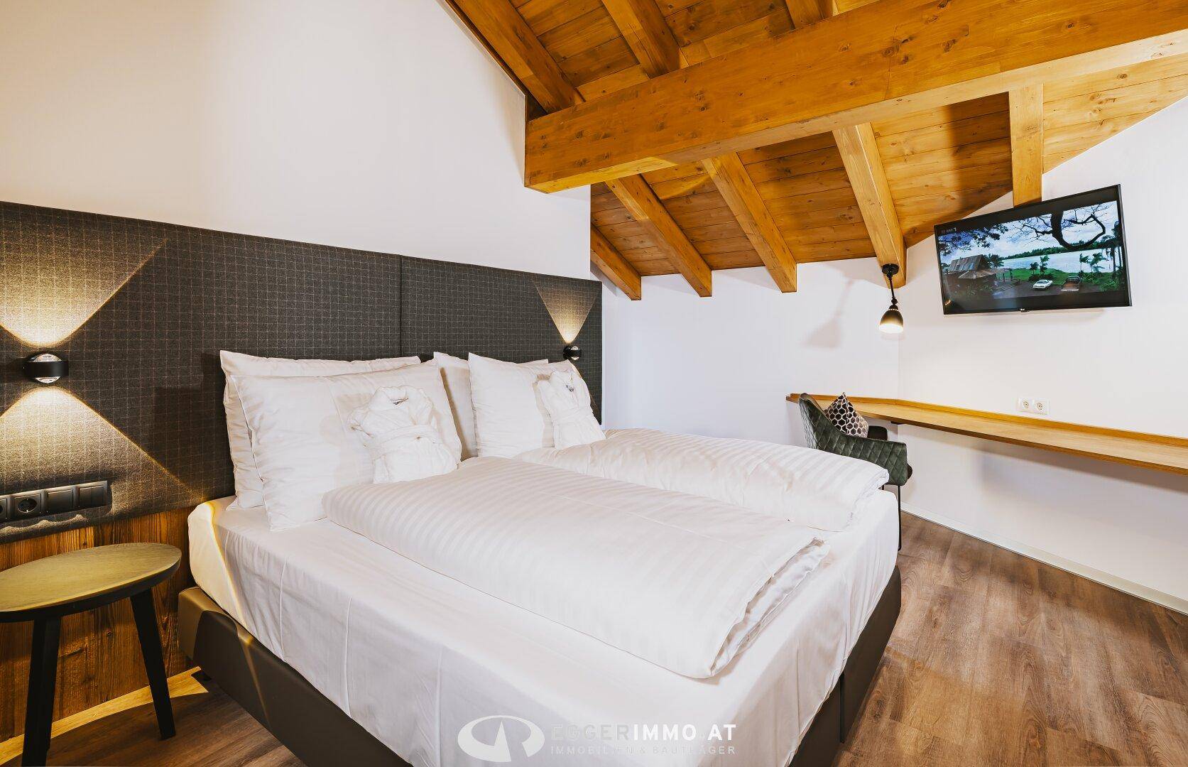 Gemütliches Schlafzimmer mit großem Bett, Holzbalkendecke und einem an der Wand montierten Fernseher.