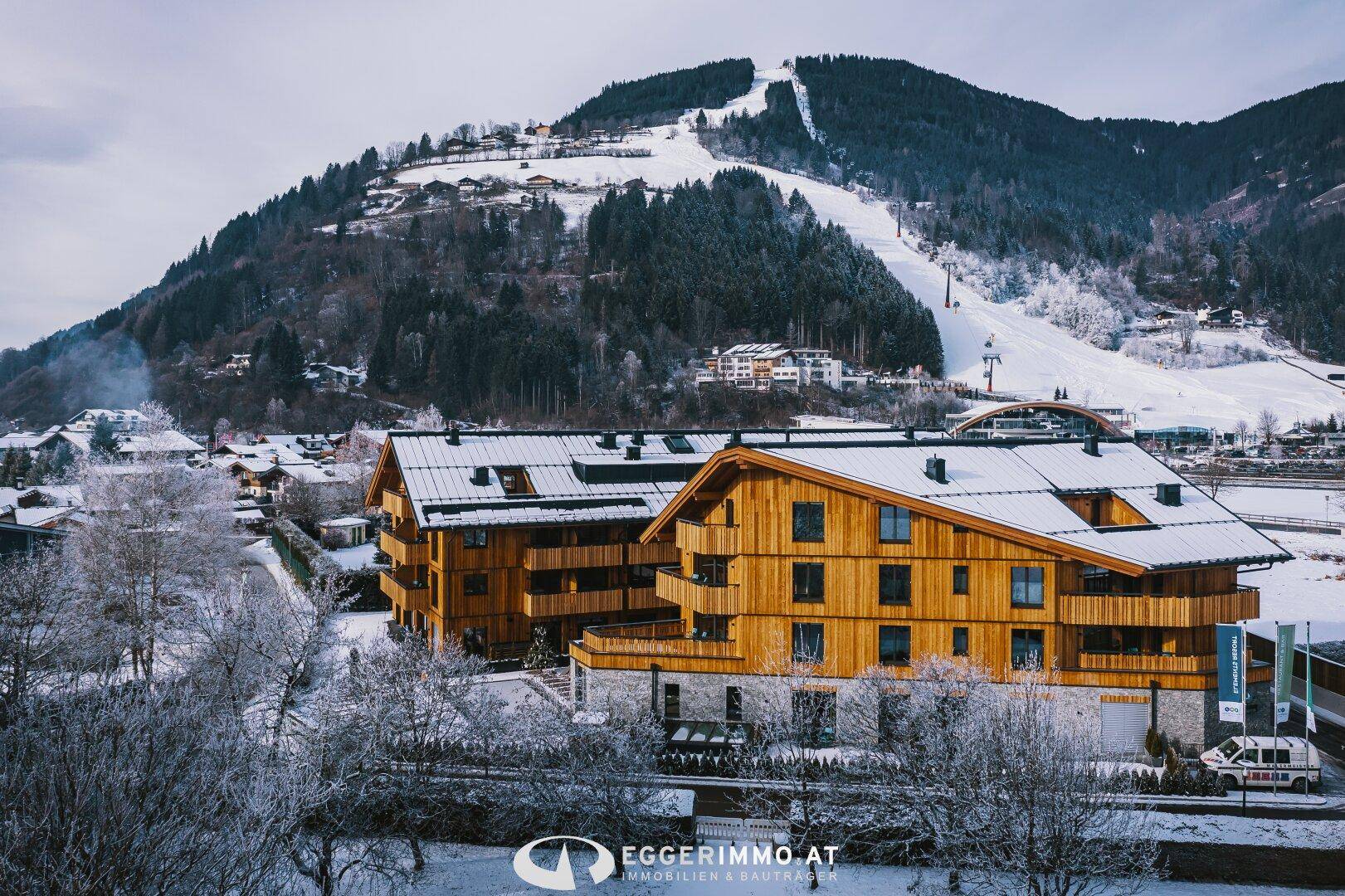 Winterlandschaft mit modernen Holzgebäuden, verschneiten Bergen und einer Skipiste im Hintergrund.