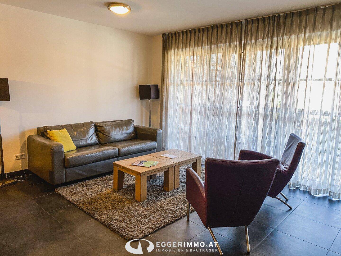 Gemütliches Wohnzimmer mit Ledersofa, Teppich und großen Fenstern mit transparenten Vorhängen.
