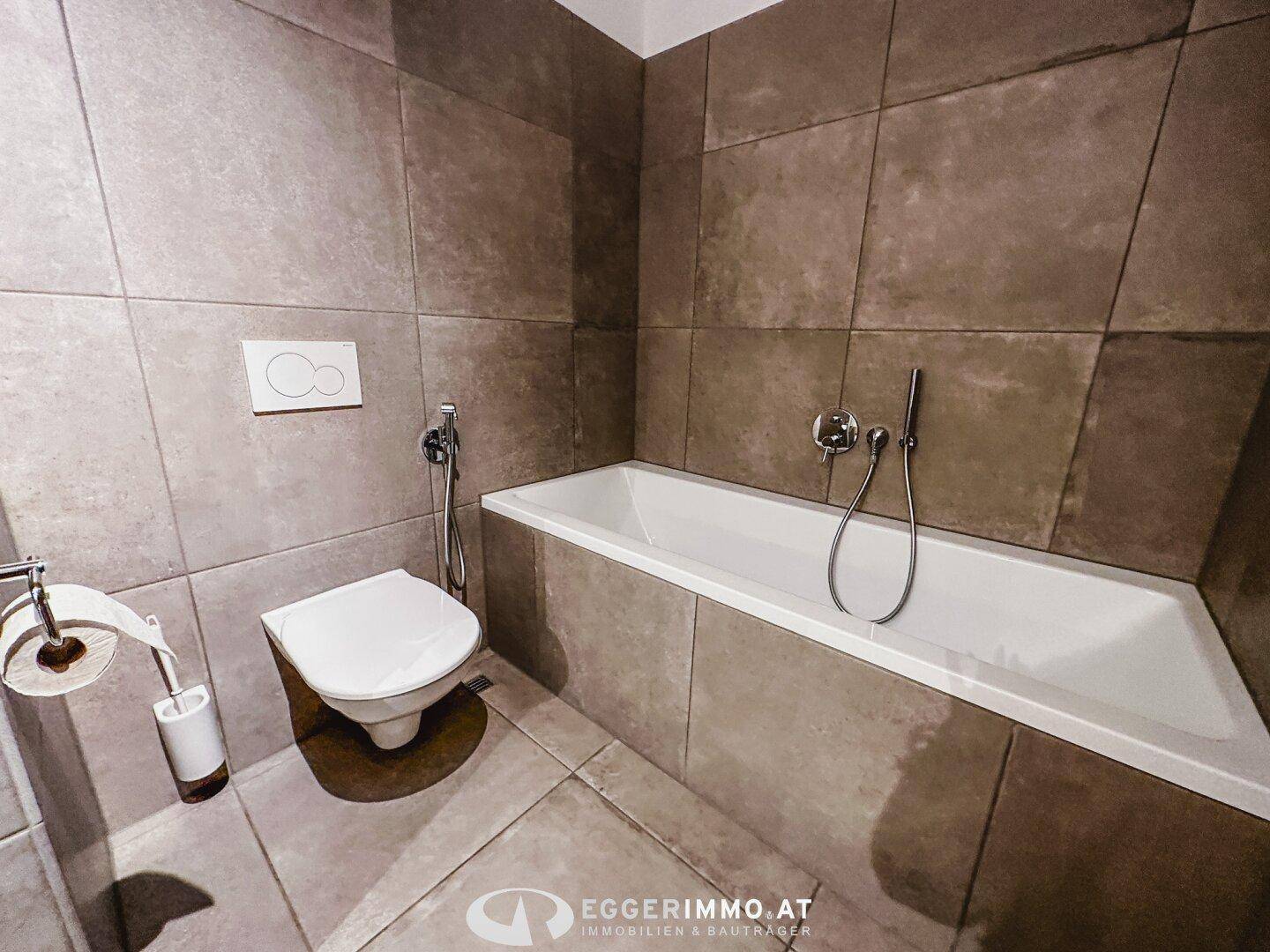 Badezimmer mit Badewanne und Toilette, ausgestattet mit großformatigen grauen Fliesen.