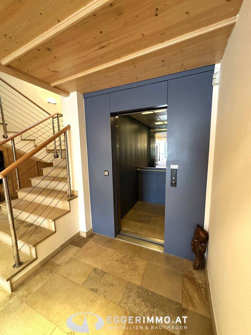 Moderner Aufzug im Flur neben einer Treppe, mit Holzdecke und Steinboden.