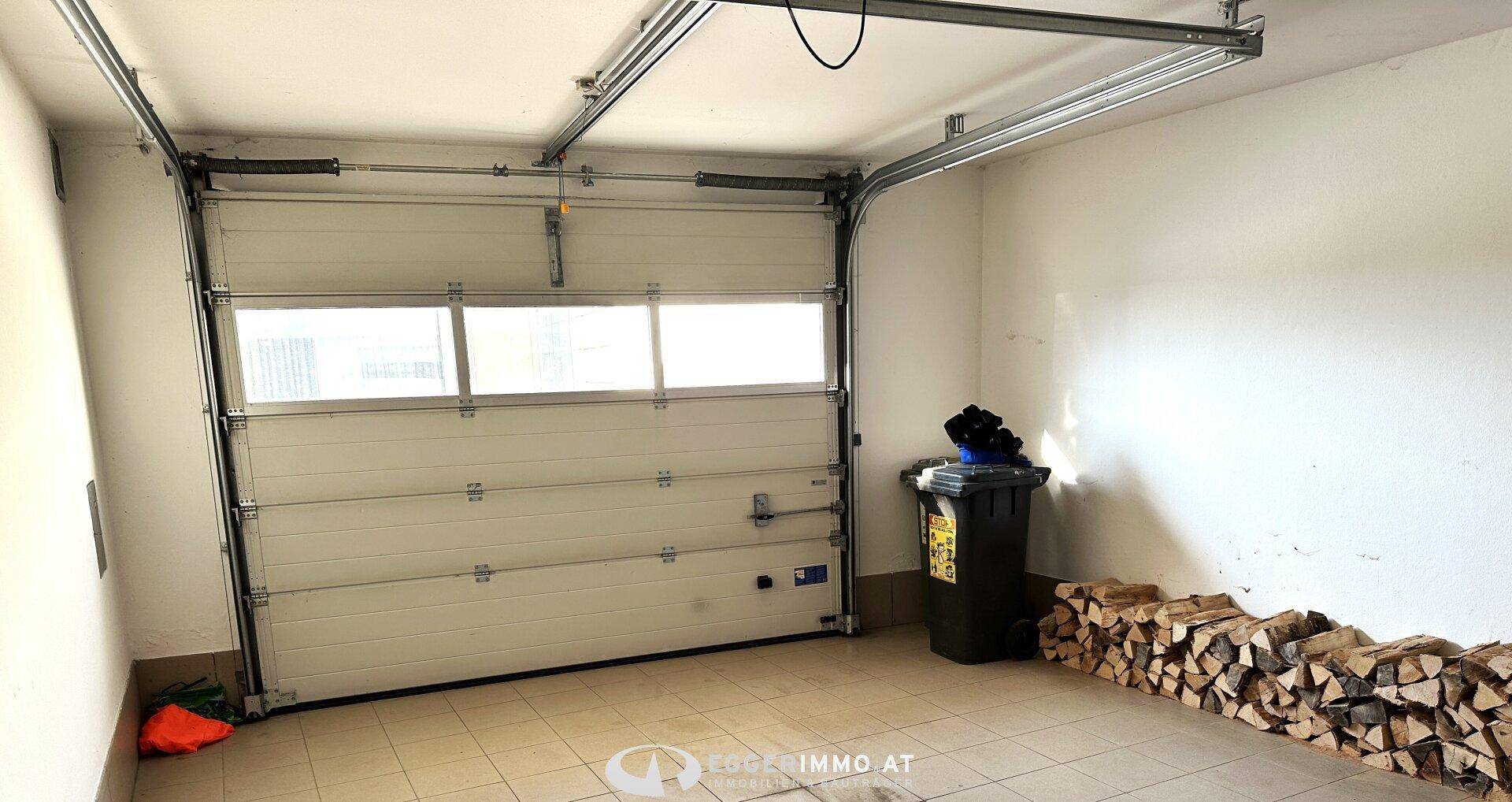 Geräumige Garage mit Sektionaltor, gefliestem Boden und gestapeltem Brennholz.